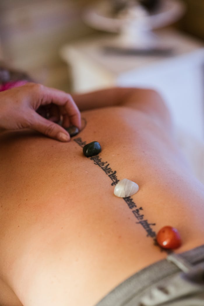 about-02 Unrecognizable female patient with tattoo and colorful stones on back getting massage from unrecognizable therapist during massage session in salon on blurred background
