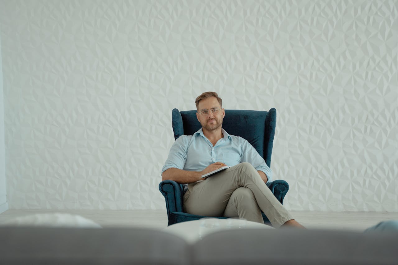 A thoughtful man sitting cross-legged in an armchair against a stylish white background.